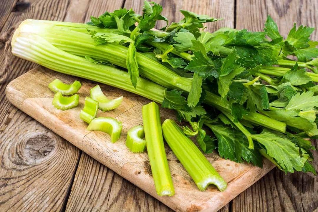 Batang seledri segar dengan daun hijau diletakkan di atas talenan kayu, sebagian telah dipotong menjadi potongan kecil.