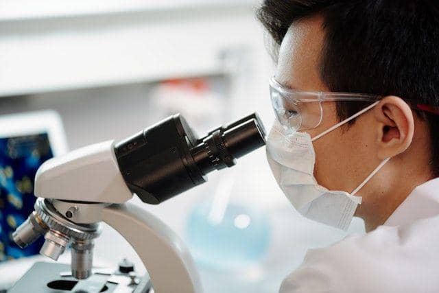 Seorang peneliti mengenakan masker dan kacamata pelindung sedang mengamati sampel melalui mikroskop di laboratorium.