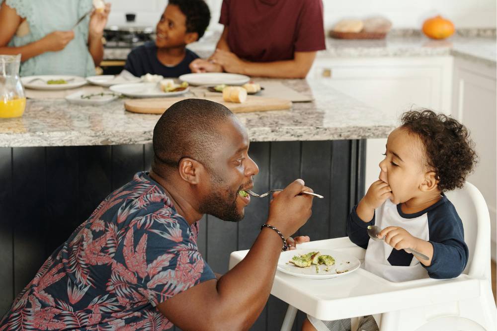 Anak disuapi ayah dengan keluarga yang makan di belakangnya