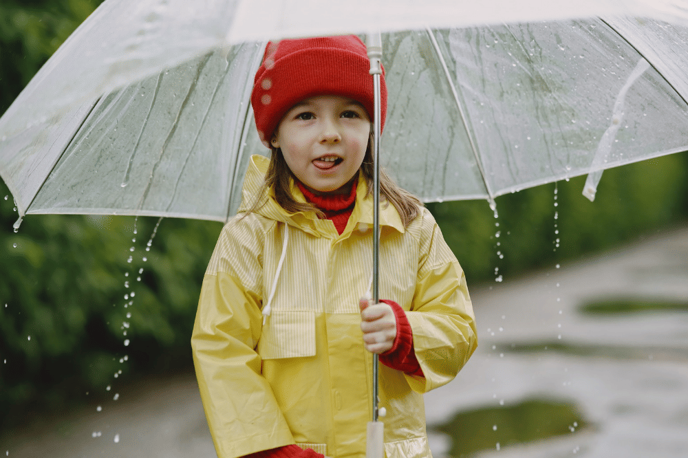 Anak perempuan sedang menggunakan payung dan jas hujan
