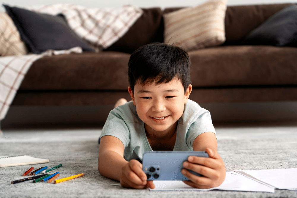 Anak laki-laki terlihat senang bermain ponsel sambil tengkurap