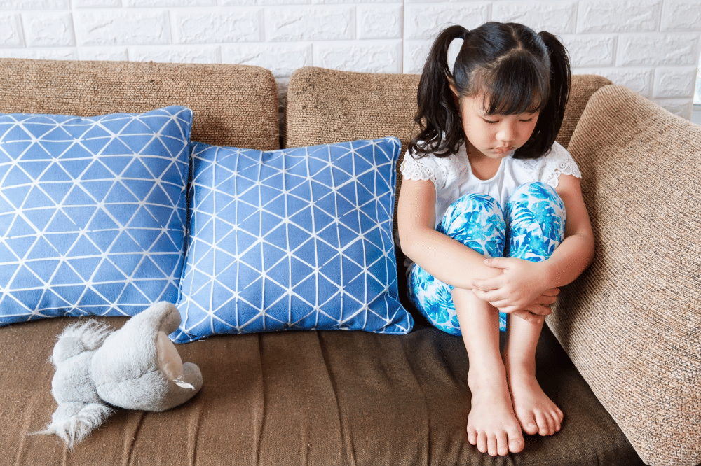 Anak perempuan duduk di atas sofa dengan menekuk kedua kaki dan terlihat murung