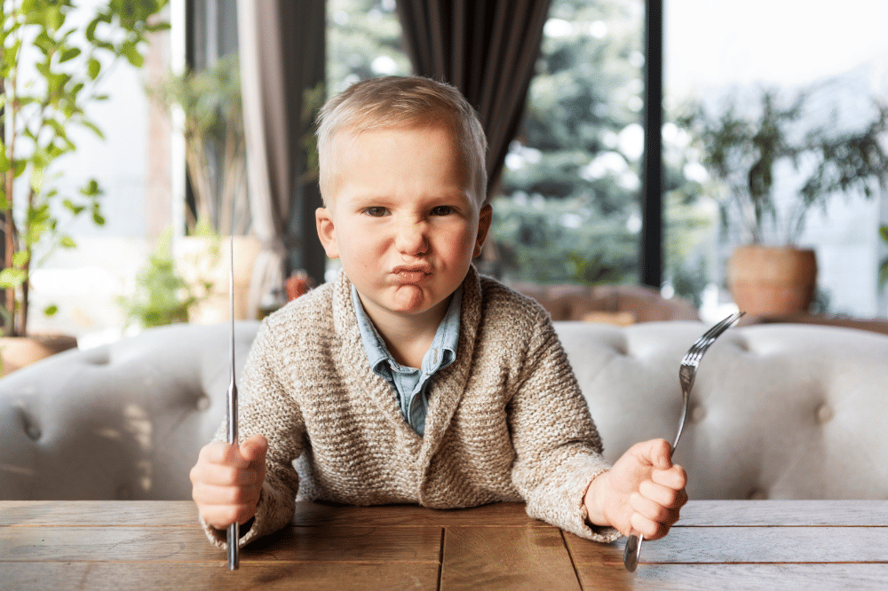 Anak laki-laki kesal sambil memegang pisau makan dan garpu karna makanannya belum siap