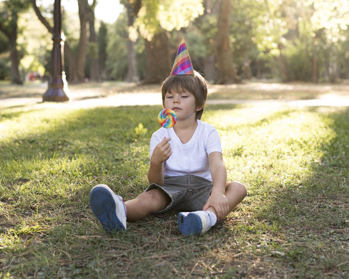 Anak laki-laki sedang makan lolipop dsambil duduk di atas rumput taman