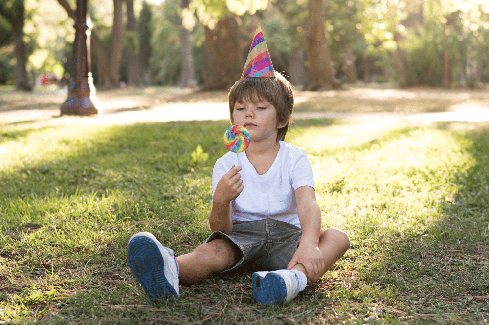 Anak laki-laki sedang makan lolipop sambil duduk di atas rumput taman