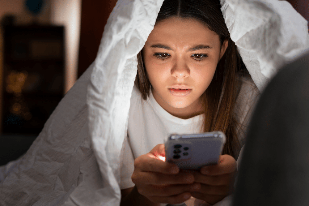 Anak perempuan sedang bermain ponsel di balik selimutnya