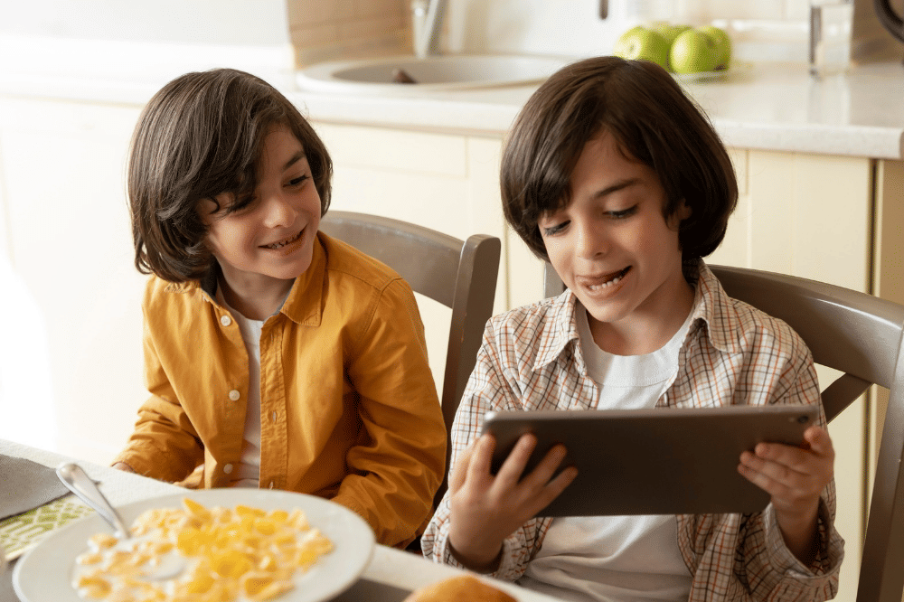 Kedua anak laki-laki tersebut sedang makan sambil bermain tablet