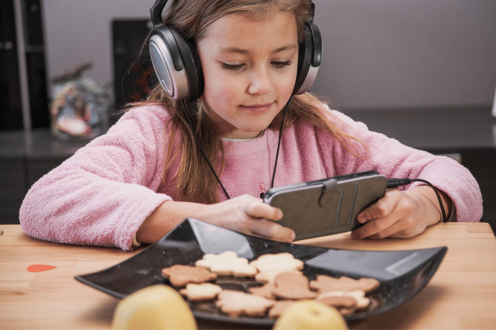 Anak perempuan sedang makan sambil menonton lewat smartphone dan headphone