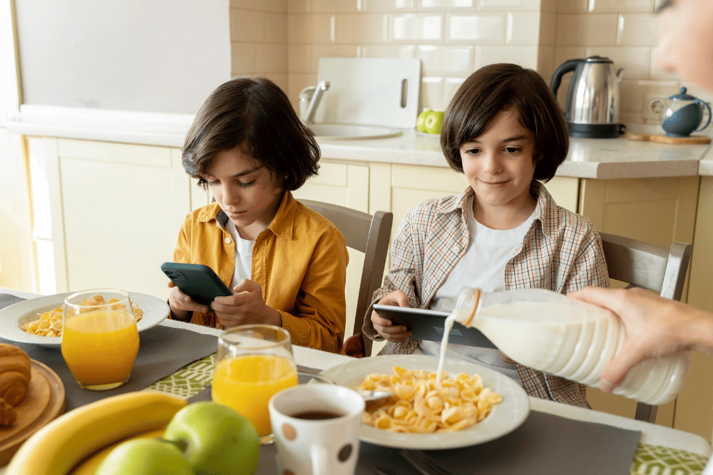 Kedua anak laki-laki tersebut sedang makan di meja sambil main gadget