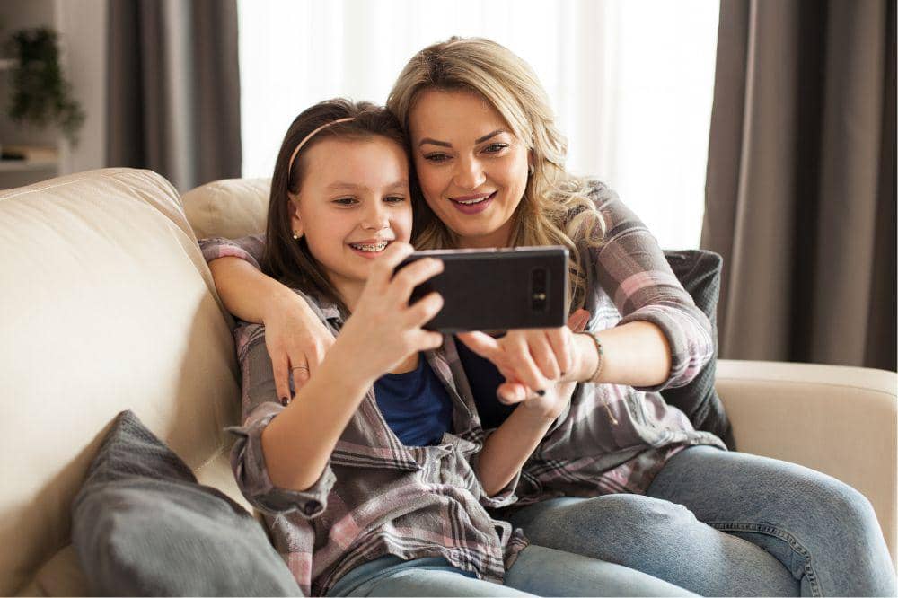 Beautiful young mother and her daughter are using a smartphone and smiling sitting on the couch in living room