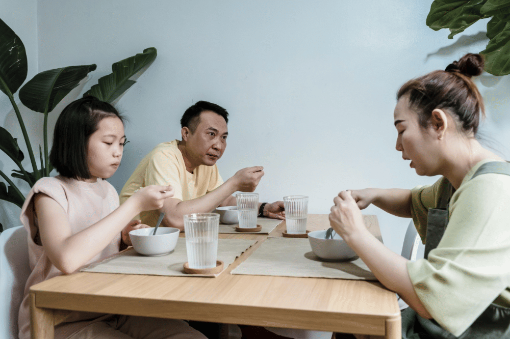 Papa, Mama, dan anak perempuannya sedang berbincang di meja makan sembari makan