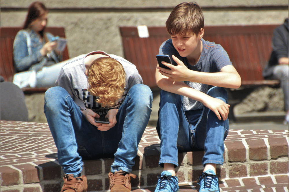 Dua remaja laki-laki duduk di pinggir jalan sembari bermain handphone 