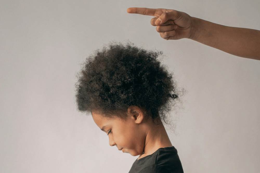 Anak kecil menunduk dengan ekspresi sedih sementara tangan orang dewasa menunjuk ke arahnya di latar belakang netral.