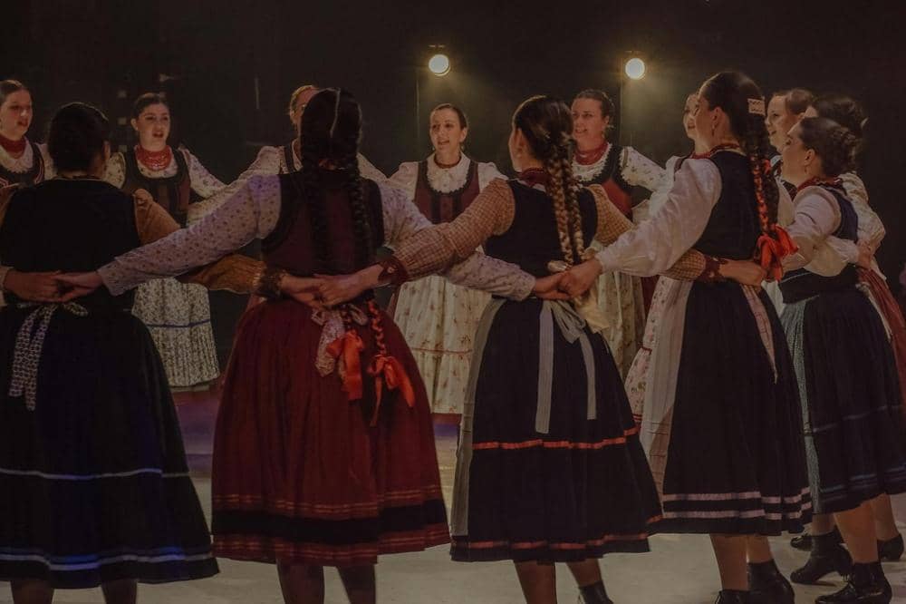 Sekelompok wanita mengenakan pakaian tradisional berwarna gelap dan terang menari dalam lingkaran di atas panggung dengan pencahayaan hangat.