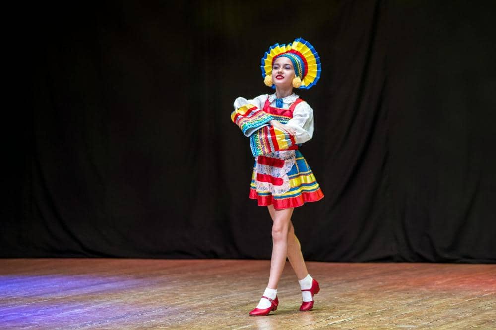 Penari mengenakan kostum tradisional berwarna cerah dengan rok pendek dan hiasan kepala besar sedang tampil di atas panggung kayu.