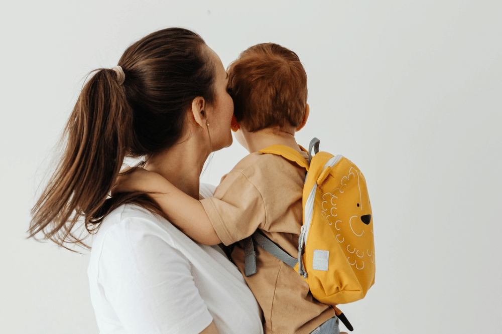 Seorang wanita memeluk anak kecil yang mengenakan ransel kuning bergambar wajah hewan di latar belakang putih polos.