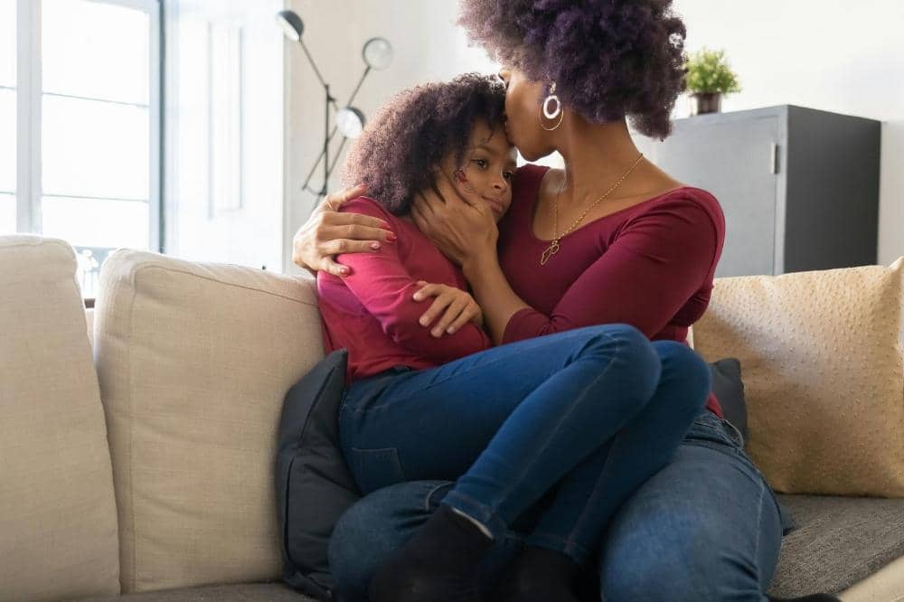 Seorang ibu memeluk dan mencium anak perempuannya yang tampak sedih di sofa ruang tamu dengan suasana hangat dan lembut.