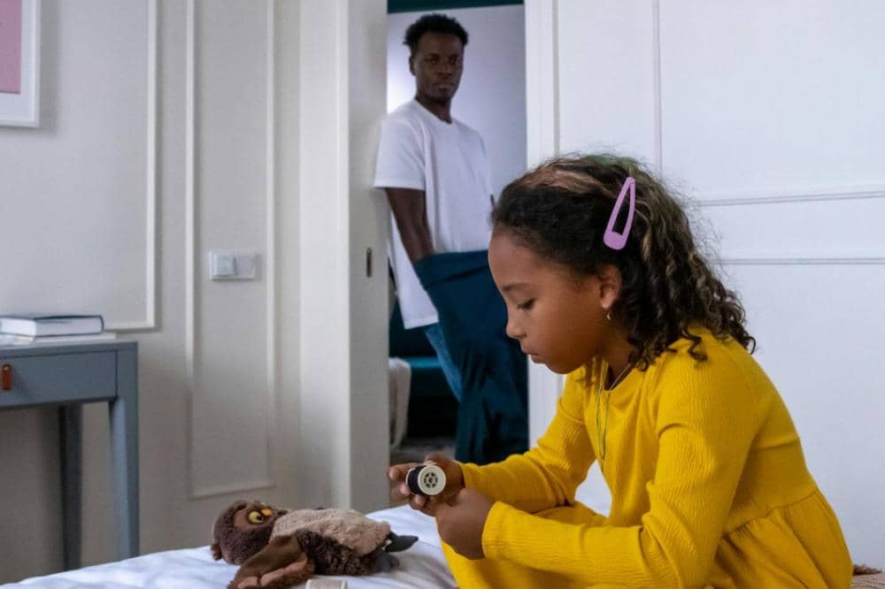 Seorang anak perempuan mengenakan gaun kuning duduk di tepi tempat tidur sambil memegang mainan