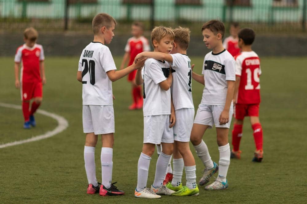 Anak-anak mengenakan seragam sepak bola putih berpelukan di lapangan hijau setelah pertandingan melawan tim berbaju merah.