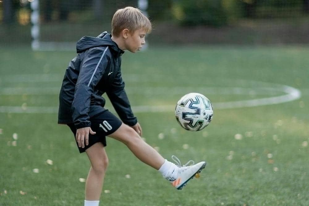 Anak laki-laki mengenakan jaket hitam dan celana pendek berlatih mengontrol bola di lapangan sepak bola hijau.