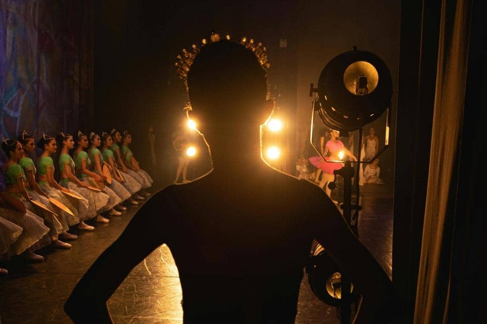 Penari balet berdiri di belakang panggung menunggu giliran tampil, sementara penari lain beraksi di bawah sorotan lampu teater.
