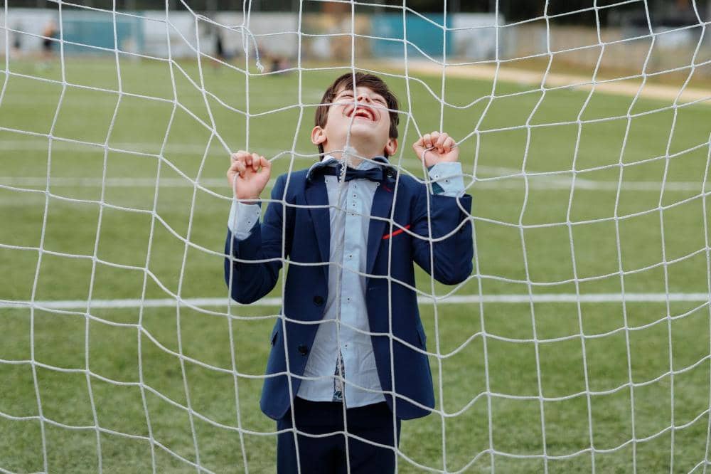 Seorang anak laki-laki mengenakan setelan jas biru berdiri di belakang jaring gawang di lapangan hijau sambil tersenyum lebar.