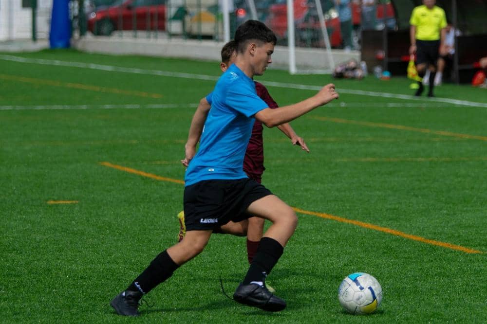 Seorang anak mengenakan seragam biru sedang menggiring bola di lapangan sepak bola hijau saat pertandingan berlangsung.