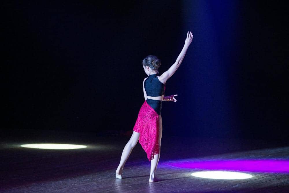 Penari wanita mengenakan kostum hitam dan merah menari di atas panggung dengan pencahayaan sorotan di sekitarnya.