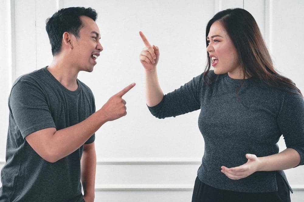 Seorang pria dan wanita sedang berdebat dengan ekspresi marah sambil saling menunjuk di dalam ruangan bercat putih.