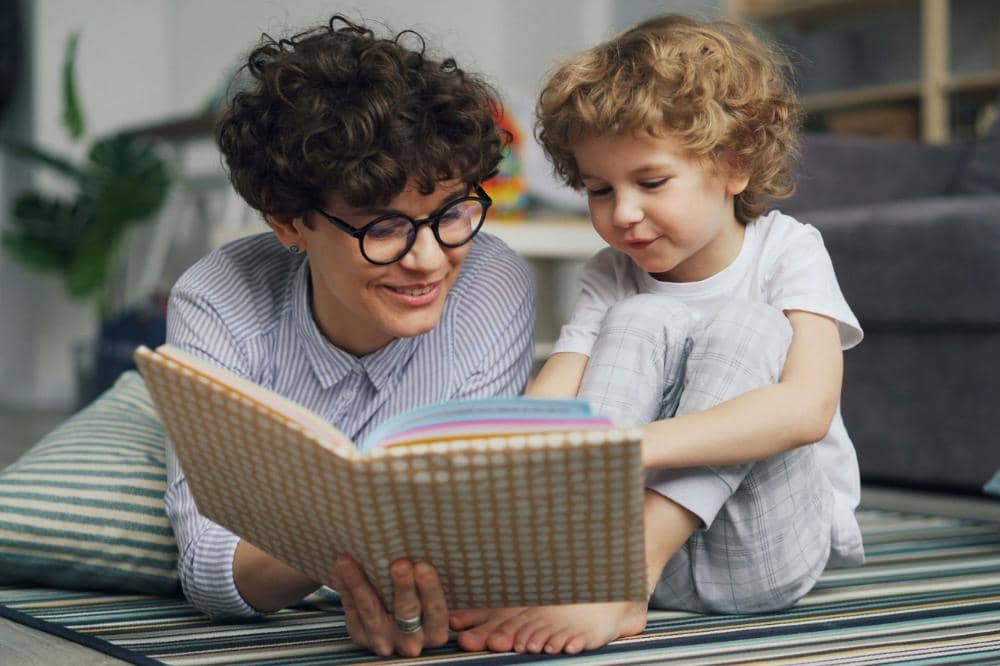 Seorang orang berkacamata tersenyum sambil membaca buku bersama anak