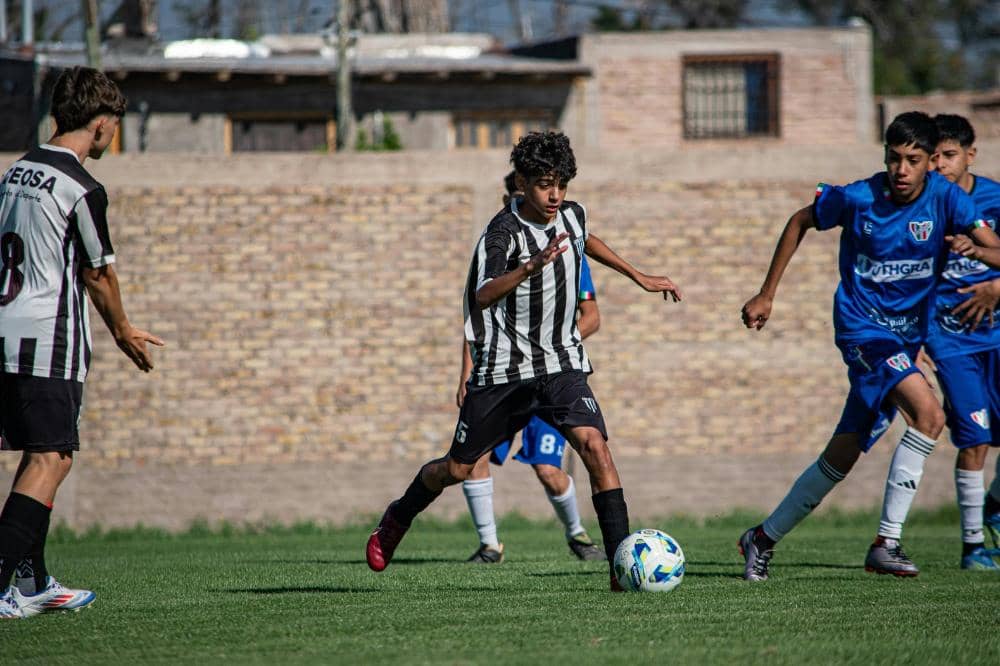 Pemain sepak bola muda mengenakan seragam hitam putih menggiring bola di lapangan hijau saat pertandingan melawan tim berseragam biru.