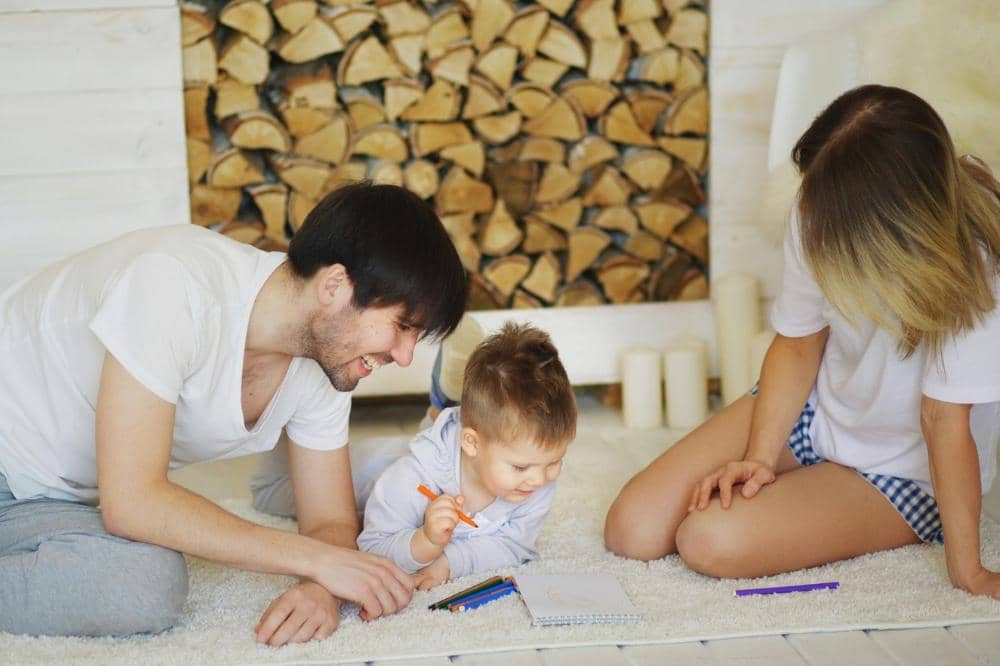 Seorang Mama dan Papa duduk di lantai bersama anak yang sedang menggambar dengan pensil warna