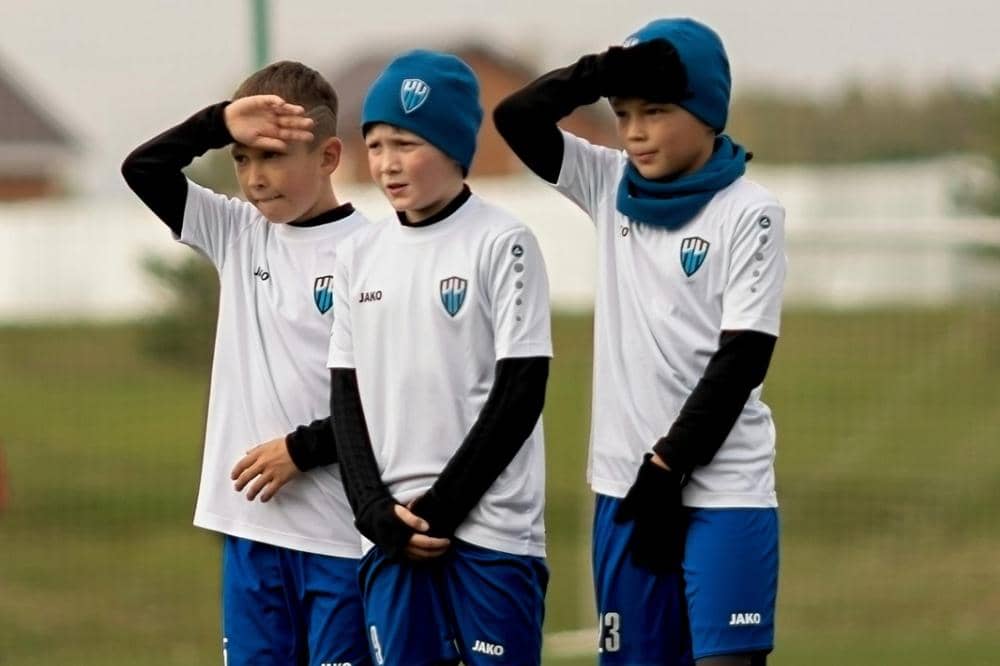 Tiga anak laki-laki mengenakan seragam sepak bola putih dan biru berdiri di lapangan sambil menutupi mata dari sinar matahari.