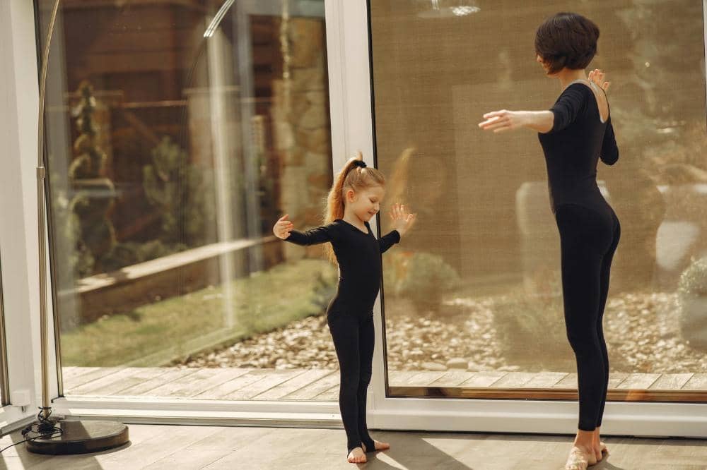 Seorang wanita dan anak kecil mengenakan pakaian senam hitam melakukan gerakan tari bersama di ruangan dengan dinding kaca besar.