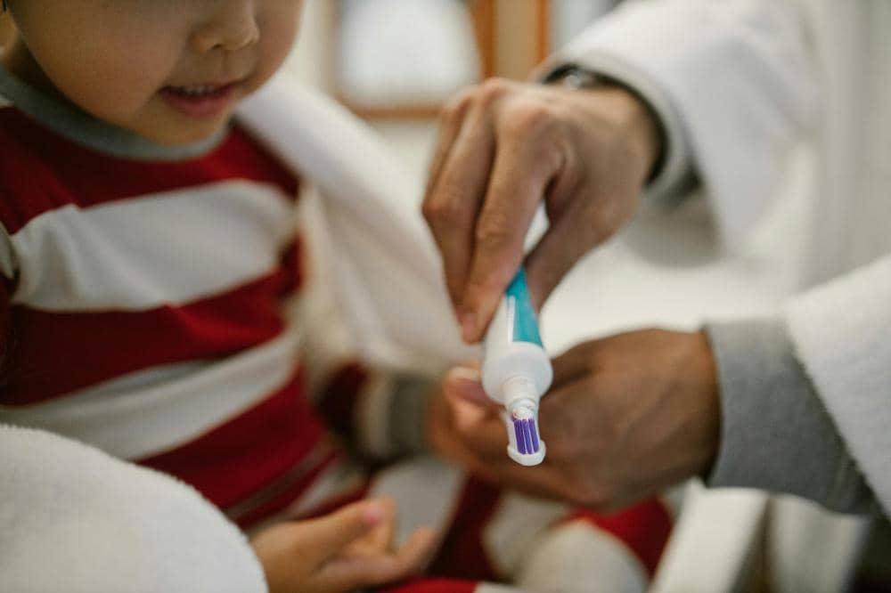 Anak kecil sedang dibantu orang dewasa mengoleskan pasta gigi ke sikat gigi.