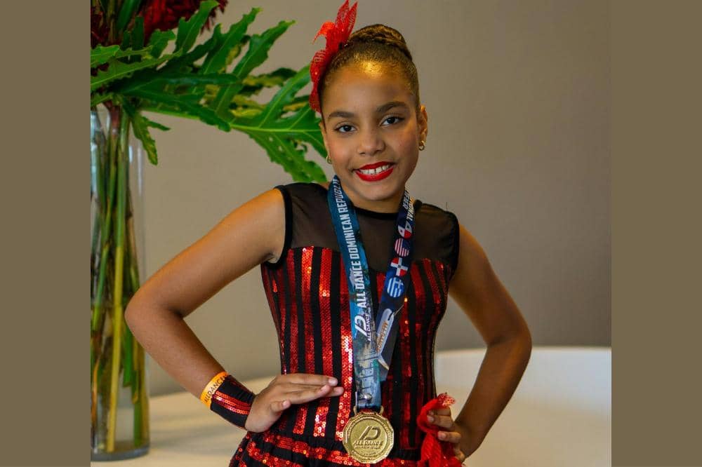 Seorang gadis muda mengenakan kostum tari merah hitam berkilau dengan medali emas di lehernya dan senyum percaya diri di wajahnya.