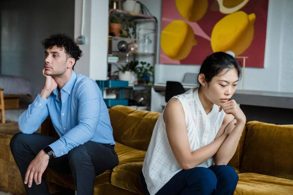 Seorang pria dan wanita duduk di sofa dengan ekspresi murung, tampak sedang berpikir atau menghadapi masalah hubungan di ruang tamu.