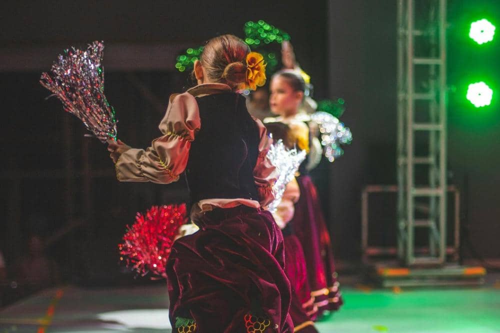 Penari mengenakan kostum tradisional berwarna merah marun dan krem dengan hiasan bunga kuning di rambut, menari di atas panggung dengan pencahayaan hijau.