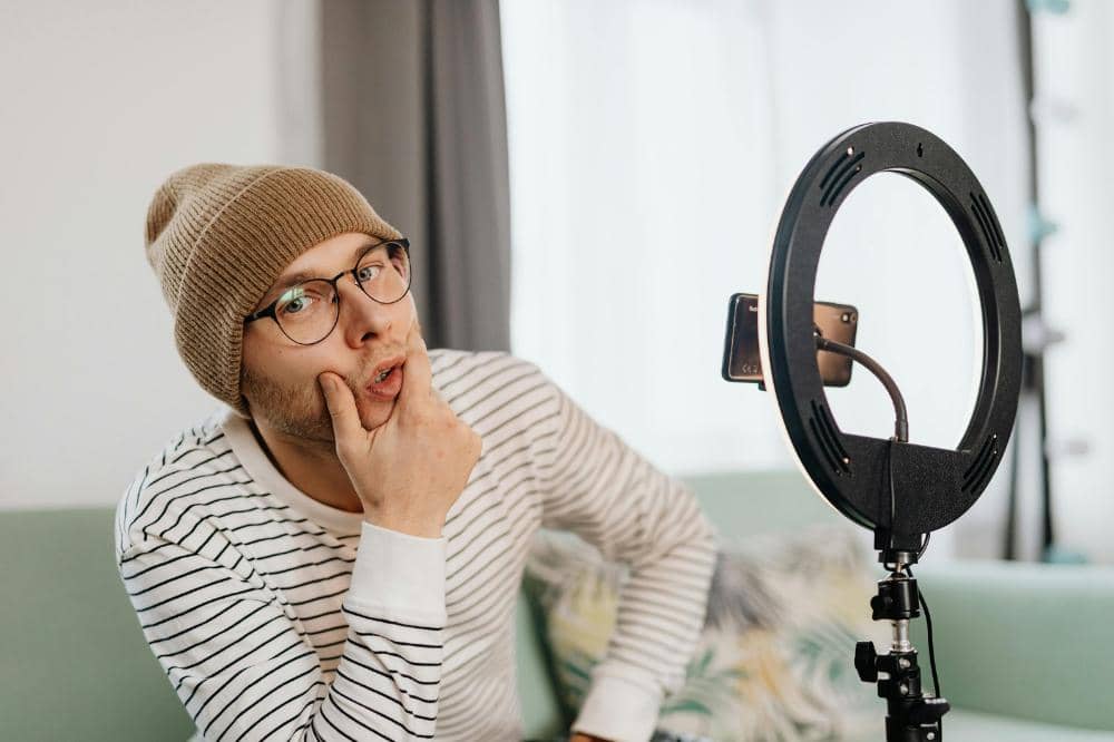 Seorang pria mengenakan topi rajut dan kacamata sedang merekam video menggunakan ponsel dan lampu ring di ruang tamu.
