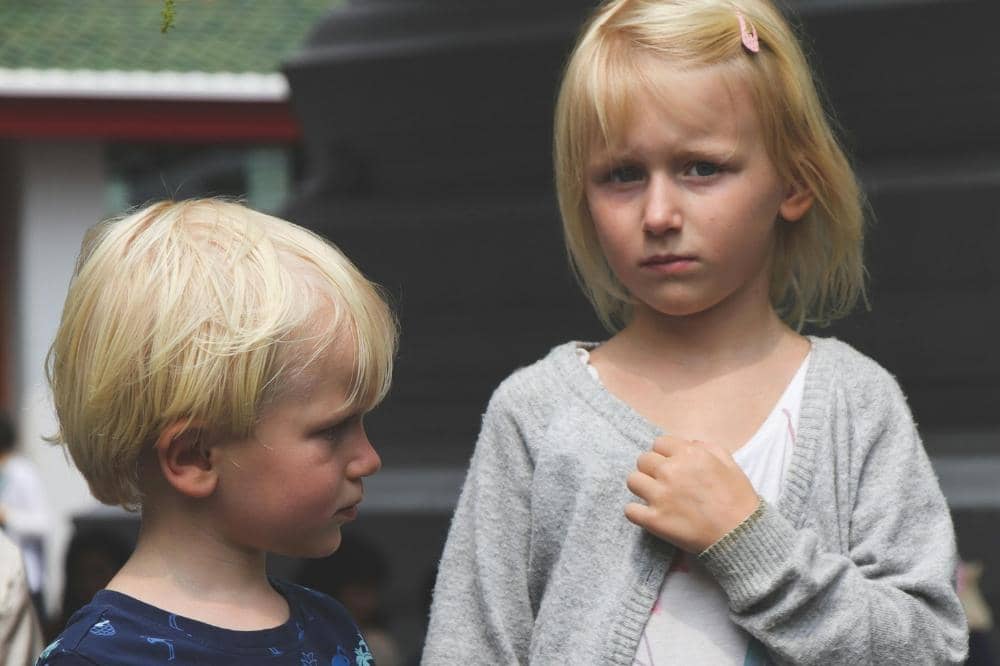 Dua anak berambut pirang berdiri di luar ruangan, satu anak perempuan mengenakan kardigan abu-abu dan anak laki-laki menatapnya.
