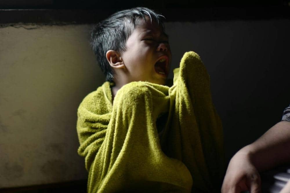 Seorang anak kecil menangis dengan tubuh dibungkus handuk berwarna hijau di dalam ruangan dengan pencahayaan redup.
