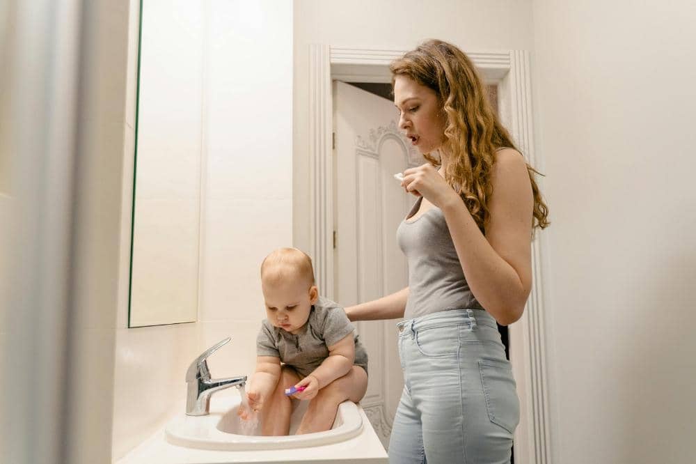 Seorang wanita berdiri di kamar mandi sambil menemani bayi yang duduk di wastafel dan bermain air di bawah keran.