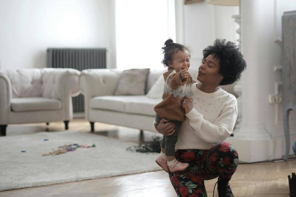 Seorang wanita berjongkok sambil menenangkan anak kecil yang menangis di ruang tamu dengan sofa berwarna terang dan karpet putih.