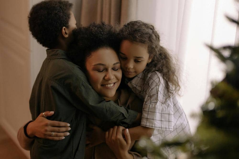 Seorang wanita tersenyum sambil memeluk dua anak di dalam ruangan hangat dengan cahaya lembut dari jendela di belakang mereka.