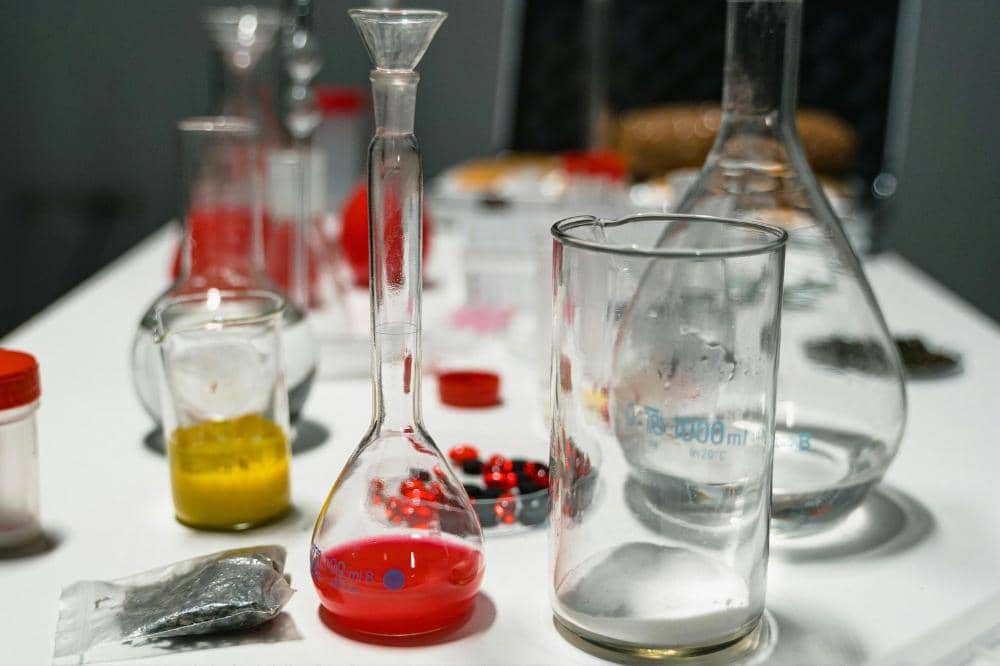 Peralatan laboratorium kimia dengan cairan berwarna merah dan kuning di dalam gelas ukur serta labu kaca di atas meja putih.