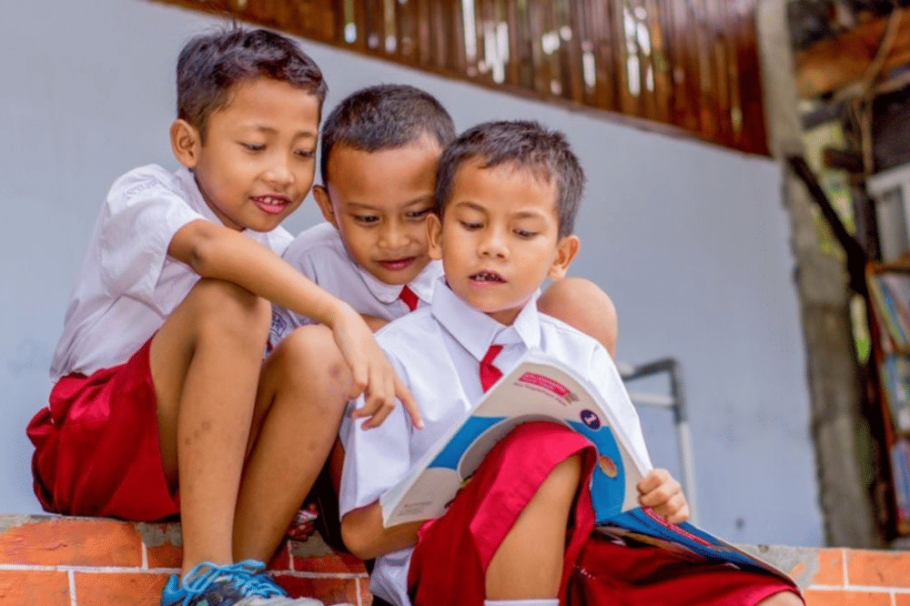 Tiga anak sekolah dasar sedang duduk sambil membaca buku.