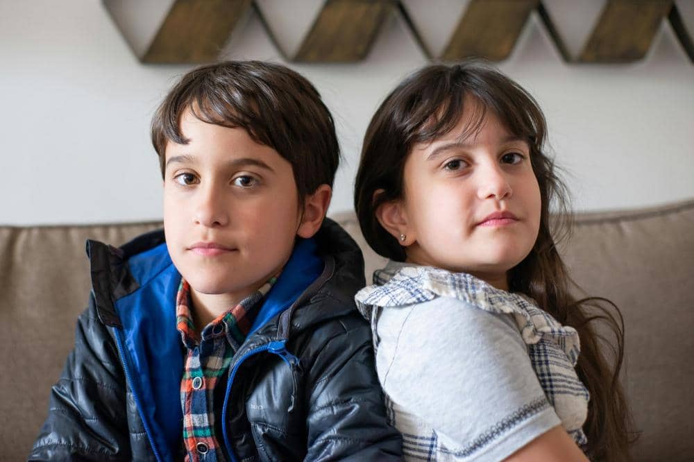 Dua anak duduk bersandar punggung di sofa, seorang anak laki-laki mengenakan jaket biru dan anak perempuan memakai kemeja kotak-kotak.