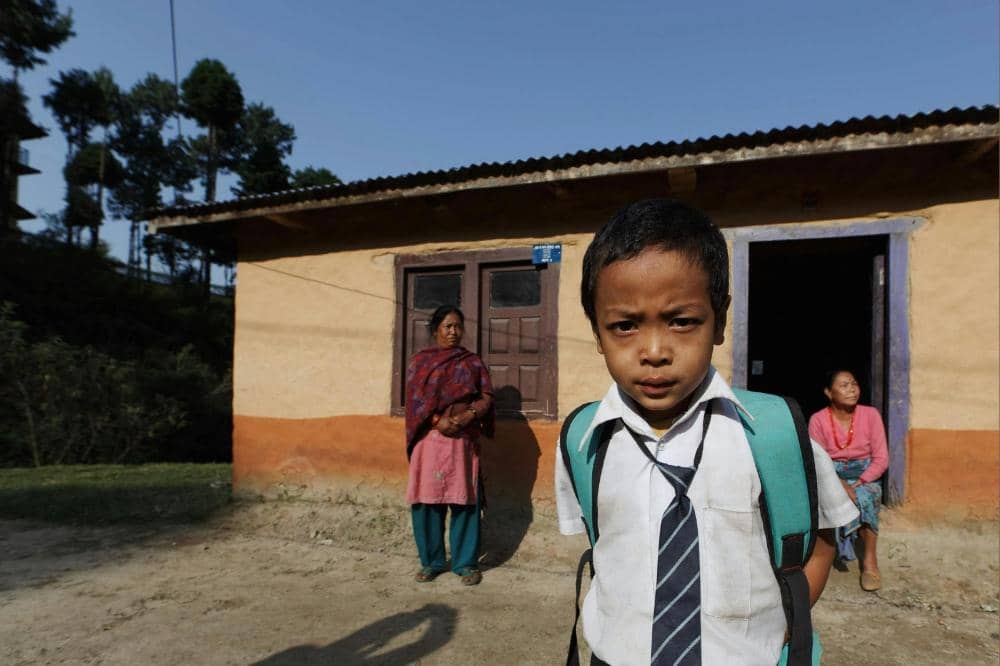 Seorang anak laki-laki mengenakan seragam sekolah dan ransel berdiri di depan rumah sederhana dengan dua wanita di latar belakang.