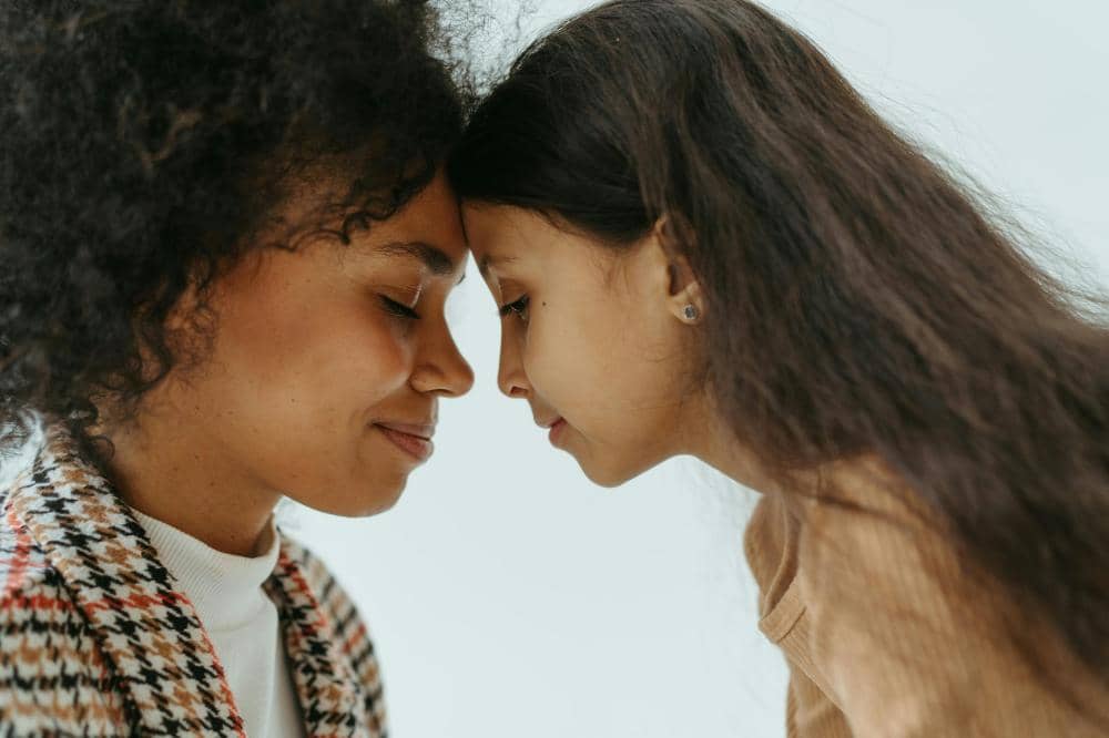 Seorang wanita dan anak perempuan saling menempelkan dahi sambil menutup mata, menunjukkan kehangatan dan kasih sayang.