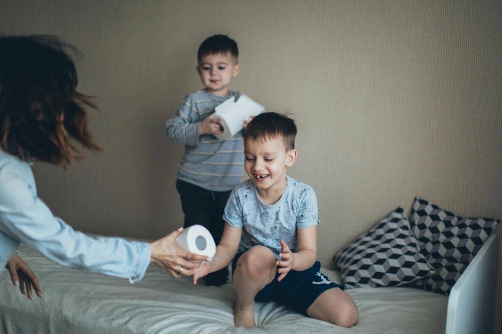 Dua anak laki-laki bermain di kamar sambil memegang gulungan tisu, dengan seorang dewasa ikut berinteraksi di tepi tempat tidur.